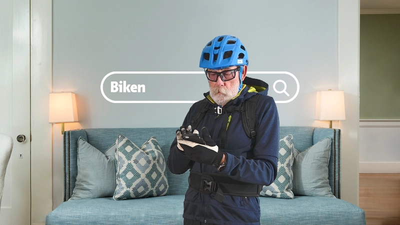 Un homme âgé portant un casque de vélo se tient dans son salon et regarde son téléphone portable